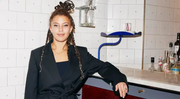 Matilda Jackson, med flätor uppsatta i en knut, lutar sig mot en bardisk med flaskor i bakgrunden.