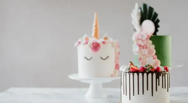 En förtrollande rosa tårta i enhörningsstil, bredvid en grön tårta med blommor och en vit tårta med chokladdryp och bär.