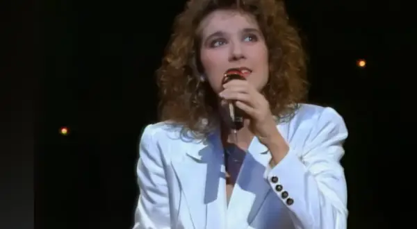 Celine Dion w białej marynarce śpiewa do mikrofonu na ciemnej scenie.