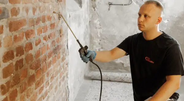 Mann sprüht mit Gerät gegen Feuchtigkeit im Keller. Ziegelwand wird behandelt.