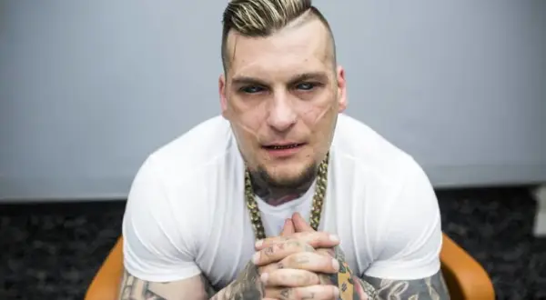 Popek kiedys, z charakterystycznymi bliznami na twarzy i tatuażami, w białej koszulce i złotym łańcuchu, siedzi na krześle.