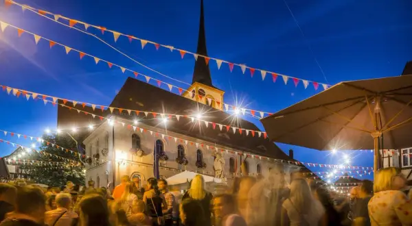Bunte Girlanden schmücken den Abend bei Weinfeste Franken. Menschen feiern vor einem beleuchteten Gebäude mit Kirchturm.