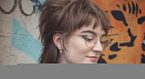 Dziewczyna w okularach z charakterystyczną, warstwową fryzurą mullet, pozuje na tle kolorowego graffiti.