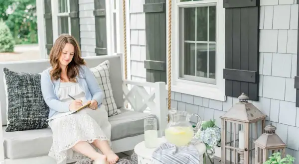 A woman embodies the coastal grandma aesthetic, writing in a notebook on a porch swing, with lemonade and hydrangeas nearby.