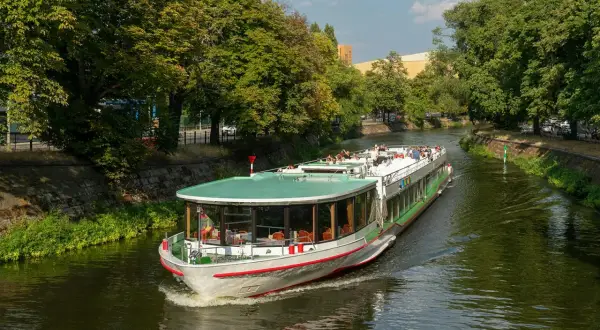 Statek wycieczkowy płynie po zielonej rzece w Berlinie, otoczony drzewami.