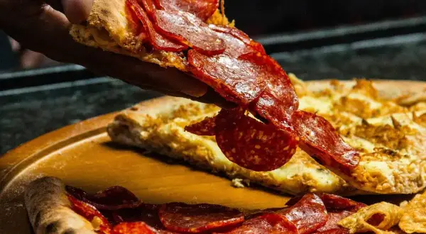 Kawałek pizzy pepperoni unoszony nad całą pizzą na drewnianej desce.