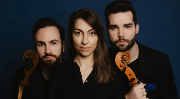Trzech muzyków, tworzących triolę, pozuje z instrumentami smyczkowymi na tle granatowym.