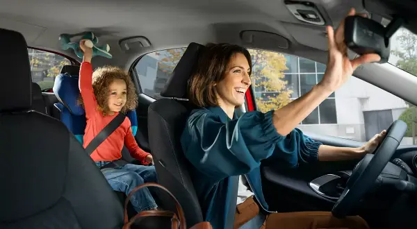 Glad mamma justerar backspegeln i sin Toyota, med barnet i bakgrunden som leker med en leksak. Med Toyota garanti kan du känna dig trygg.