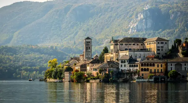A picturesque Italian village with colorful buildings and a tall bell tower sits on the edge of a calm lake, with tree-covered mountains in the background.