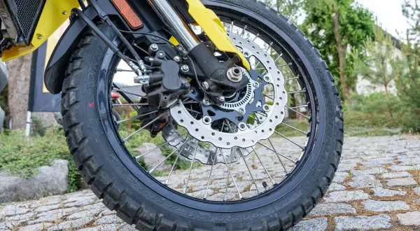 Koło motocykla z tarczą hamulcową i zaciskiem, na tle kamiennej nawierzchni i zieleni.