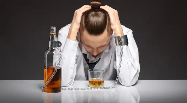 Mężczyzna w kajdanach, przykuty do butelki alkoholu i szklanki. Obraz symbolizuje uzależnienie i pokazuje, jak działa wszywka alkoholowa, jako symbol zniewolenia.
