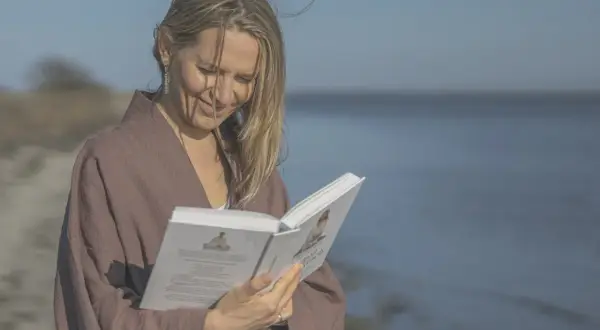 Kobieta w brązowym kimonie czyta książkę na tle morza.