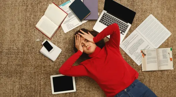 Zmęczona studentka leży na dywanie otoczona książkami, laptopem i telefonem.
