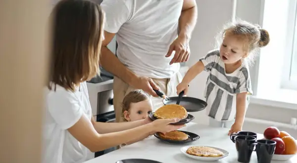 Rodzina przygotowuje placuszki dla niemowlaka 7 miesięcy. Mama podaje talerz z placuszkami, a tata i starsza córka pomagają na patelni.