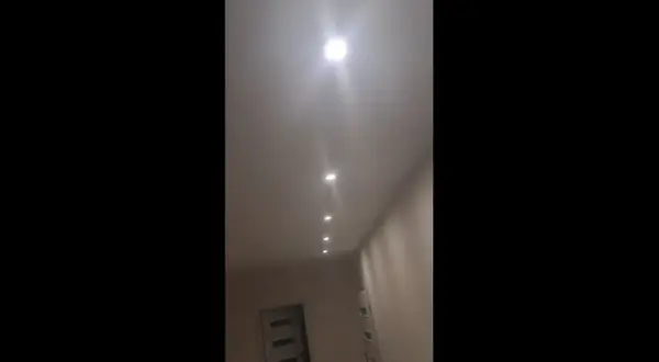 W korytarzu domu widać rząd wbudowanych w sufit lamp. Jedna z nich, najbliżej widza, świeci najjaśniej, tworząc efekt migającego światła.