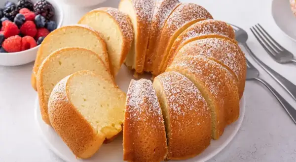Pyszna babka piaskowa, jak z przepisu babci, posypana cukrem pudrem, z owocami i widelcami obok.