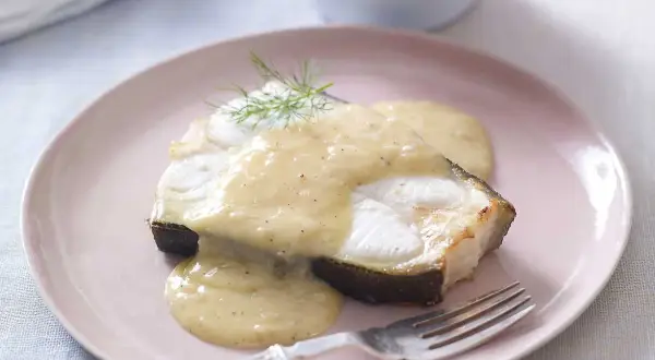 En bit fisk med en krämig beurre blanc-sås, garnerad med dill. Serveras på en ljusrosa tallrik med en gaffel.