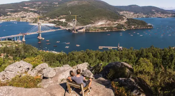 Pareja sentada en un mirador, contemplando el impresionante puente de Rande sobre el mar, con bateas de mejillones y un pueblo al fondo.
