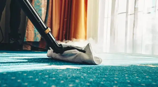Dampfreiniger reinigt einen blauen Teppich mit Muster. Dampf steigt auf, während die Düse über den Boden gleitet.