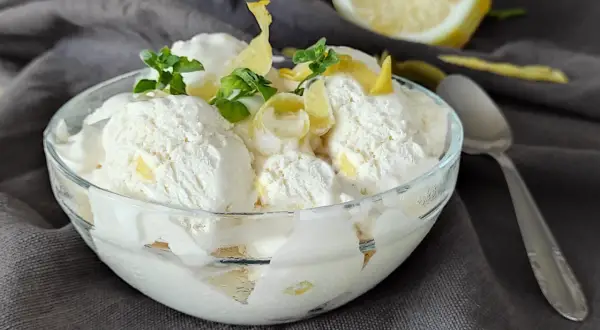Ein Glas cremiges zitronen eis, garniert mit Minze und Zitronenschale, daneben eine halbe Zitrone und ein Löffel.