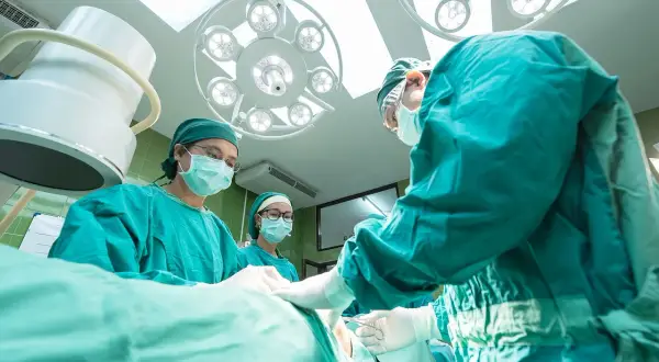 Zespół chirurgów w zielonych fartuchach pracuje pod jasnymi lampami. Obserwują pacjenta, być może analizując objawy erozji taśmy TOT.