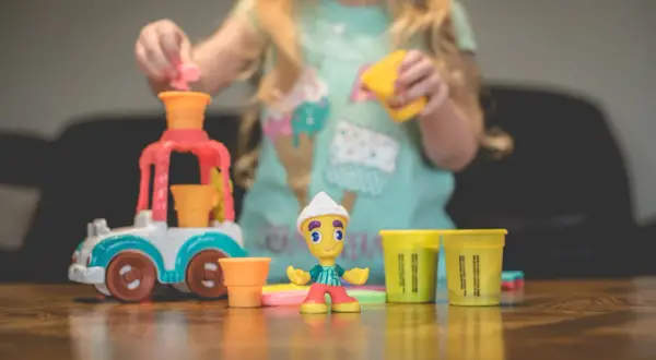 Dziecko bawi się w play doh warsztat, tworząc lodowe przysmaki. Na stole kolorowe foremki i figurka lodziarza.