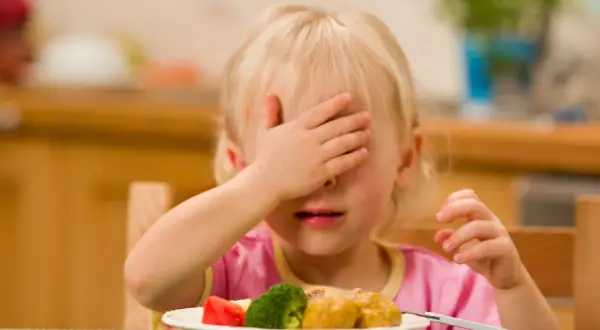 Mała dziewczynka zakrywa oczy ręką, patrząc na talerz z brokułami i klopsikami. Wygląda, jakby mówiła "je wciąż to samo".