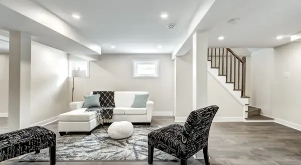 A bright, modern living area in a finished basement, featuring a white sectional, patterned rug, and a staircase leading up.
