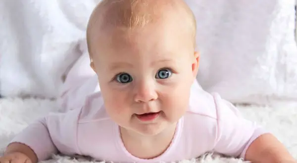 Bebé con ojos azules y cabello rubio, con un mameluco rosa. Pensando en nombres de chica ingleses como Emily o Olivia.
