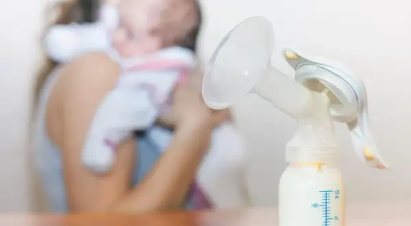 Laktator z butelką mleka, w tle mama z dzieckiem. Nawał pokarmu w której dobie to wyzwanie, ale dzięki niemu maluch ma co jeść.