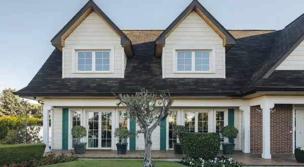 Fachada de casas Canexel con ventanas blancas, contraventanas verdes y un jardín cuidado con un olivo.