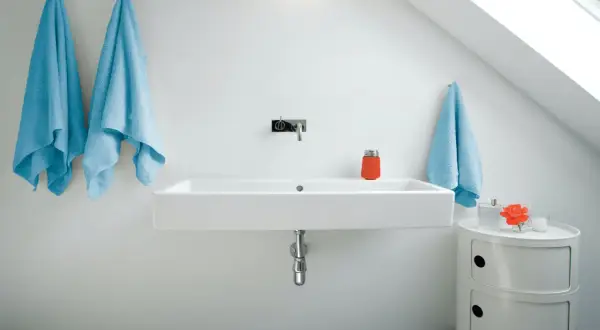 Modernt badrum med ett stilrent handfat. Två ljusblåa handdukar hänger på vänster sida, en på höger. En orange behållare och en blomma står på ett litet vitt skåp.