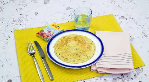 Tortilla de gambas dorada en plato blanco con borde azul, servida con cubiertos, vaso de agua y caldo de pescado.