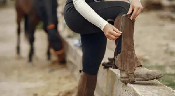 Jeździec zapina sztylpy, które chronią łydki. W tle koń pasie trawę.