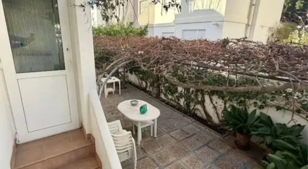 Patio con mesa y sillas bajo una pérgola cubierta de enredaderas secas. Un gran cortijo se vislumbra al fondo.