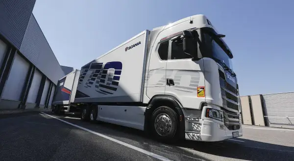 Un camion blanc Scania 770S avec une remorque, garé devant un bâtiment industriel sous un ciel bleu clair.