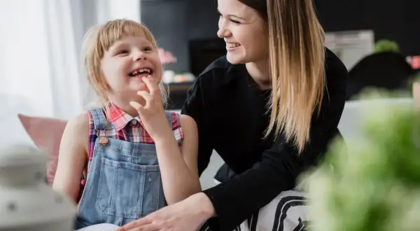 Uśmiechnięta dziewczynka z niepełnosprawnością sprzężoną bawi się z mamą, która wspiera ją w nauce.