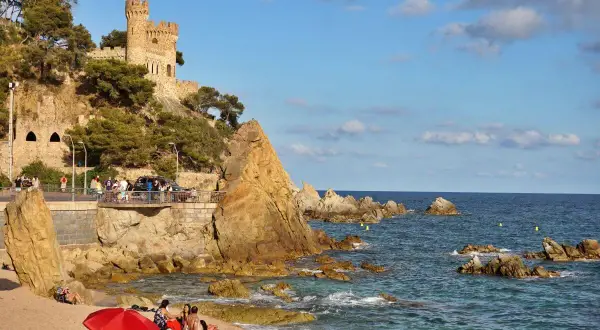 El kasteel Lloret de Mar se alza sobre una colina rocosa junto a una playa con gente disfrutando del sol y el mar azul.