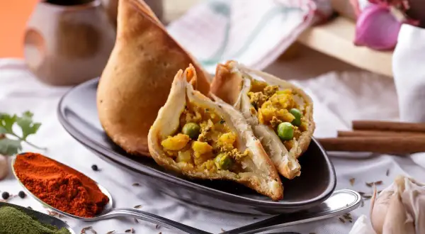 Chrupiący indyjski pierożek samosa, przekrojony na pół, ukazuje bogate nadzienie z ziemniaków i groszku. Obok łyżki z papryką i zielonymi przyprawami.