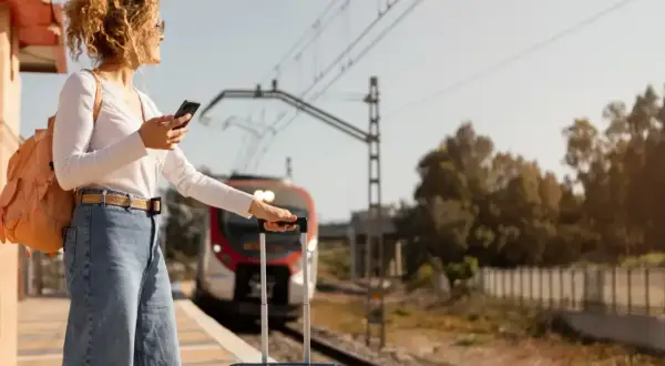Kobieta z plecakiem i walizką na peronie sprawdza telefon, zastanawiając się, ile się płaci za nadbagaż. Pociąg zbliża się do stacji.