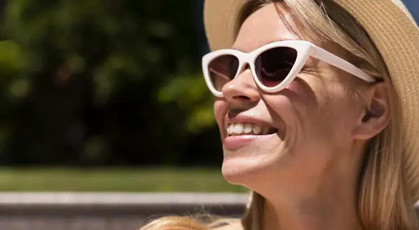 Donna sorridente con cappello di paglia e occhiali da sole, le rughe da sole che le solcano il viso.