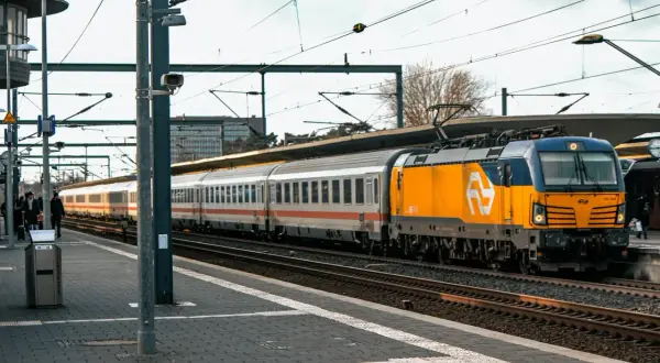 Pociąg z żółtą lokomotywą stoi na stacji, pasażerowie czekają, być może z powodu opóźnienia.