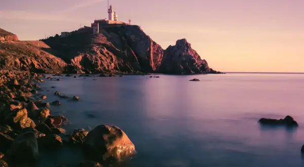 Faro en un acantilado rocoso al atardecer, con el mar tranquilo y piedras en la orilla. Parece la boca de los frailes.