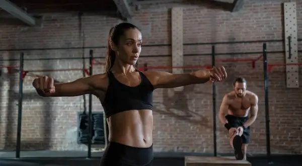 A woman performs boxing neck exercises in a gym. A man stretches in the background.