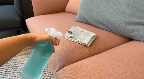 Hand sprüht Reinigungsmittel auf einen rosafarbenen Sofa-Stoff, um Wasserflecken zu entfernen. Ein Tuch liegt daneben.