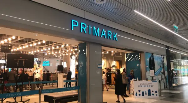 Wejście do sklepu Primark. W środku widać manekiny i ubrania. Na ladzie informacyjnej znajduje się **Primark tabela rozmiarów**.