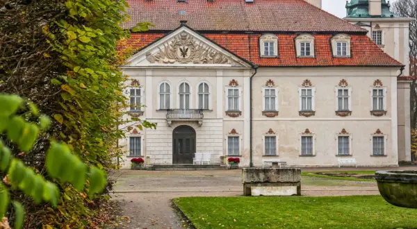 Pałac w Nieborowie z czerwoną, ceramiczną dachówką i ozdobnym frontonem. Wokół jesienne liście i bujna zieleń.