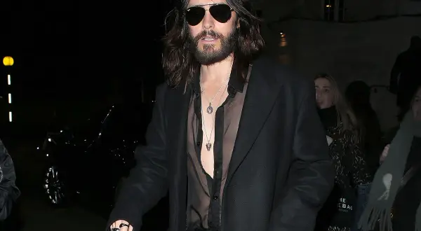Jared Leto con un audace outfit camicia nera trasparente sotto un cappotto scuro, collane d'argento e occhiali da sole.