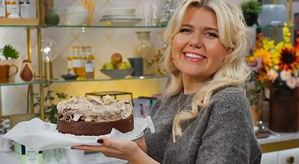 En leende kvinna håller upp en kladdig kladdkaka med fluffig topping. Bakom henne syns hyllor med köksdetaljer och blommor.