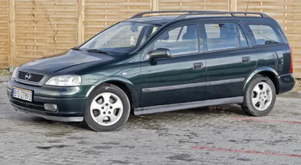 Zielony Opel Astra kombi, jeden z wielu aut w ocynku, stoi na mokrym parkingu przed drewnianym płotem.